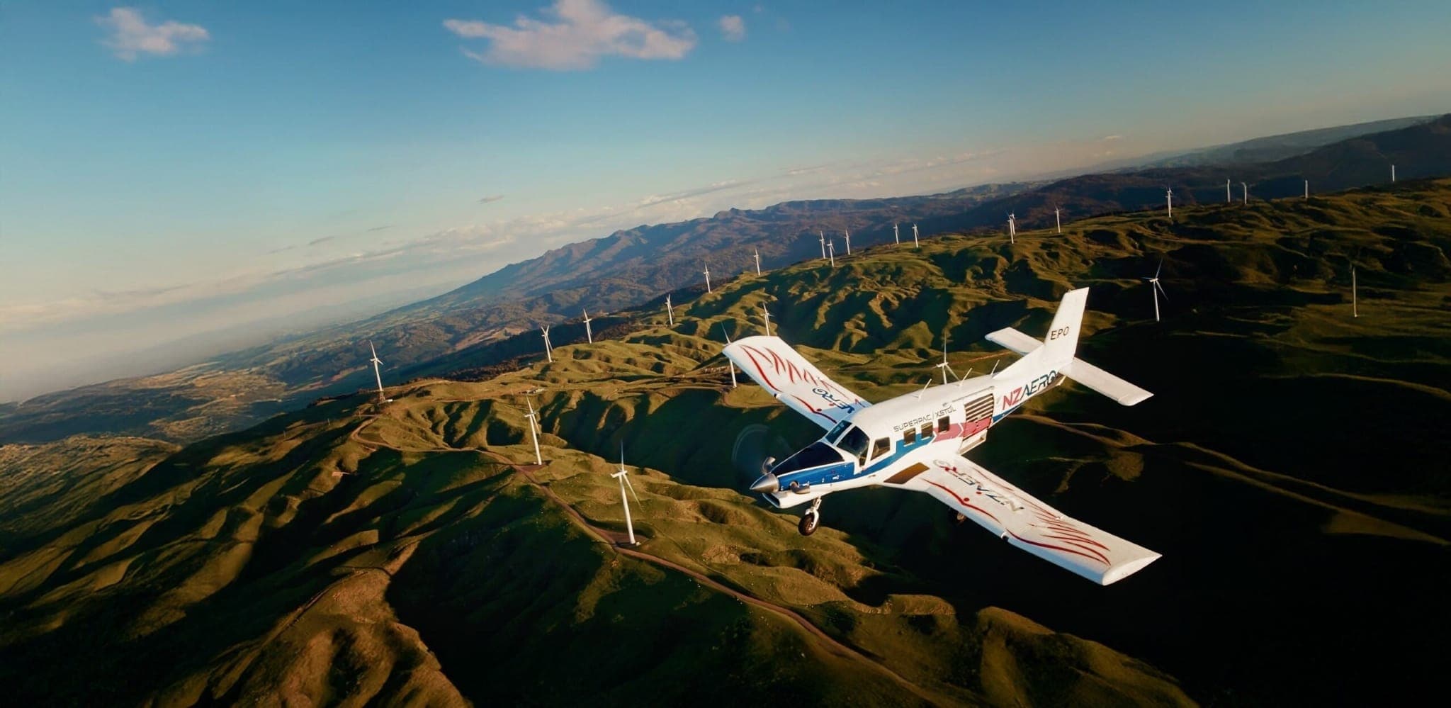 SuperPac 750XL-II in volo sopra colline verdi e turbine eoliche